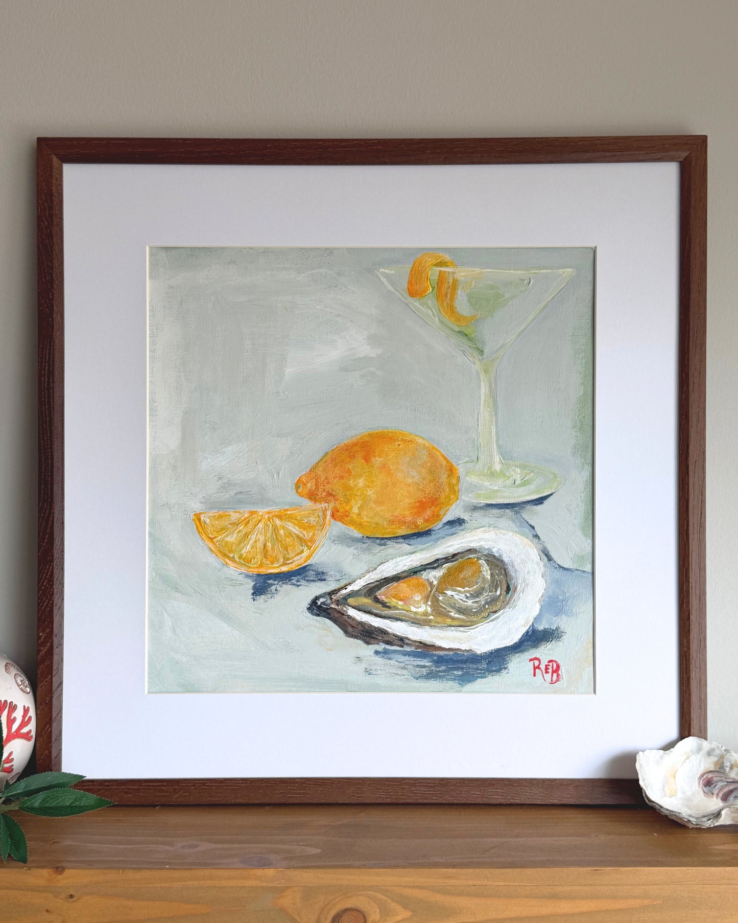 Framed painting of a cocktail glass, lemon, and oyster on a table.