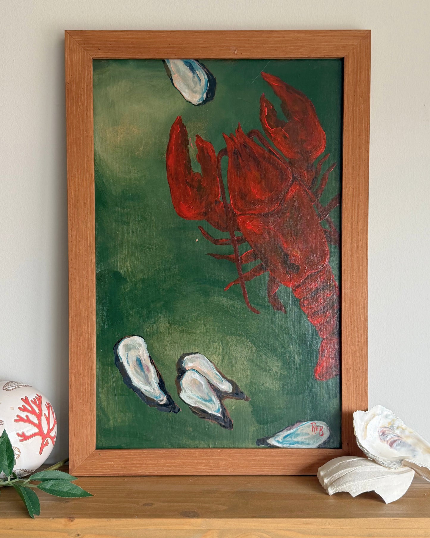 Framed painting of a red lobster on a green background with oysters, displayed on a surface.